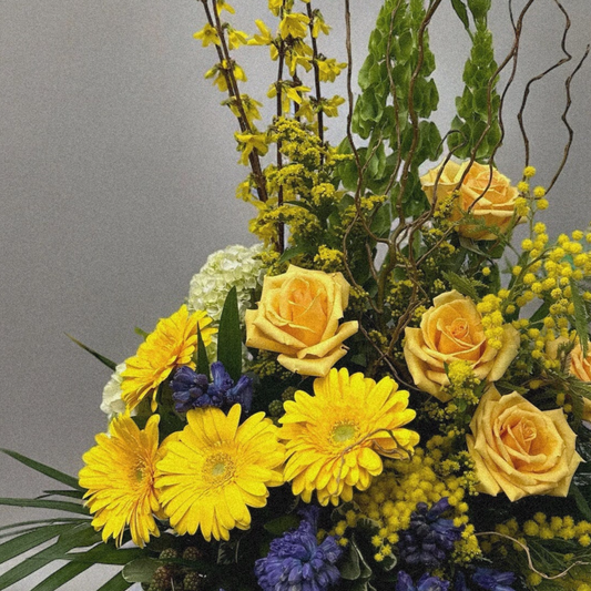 Sunny Elegance Arrangement with yellow roses, gerberas, blue hyacinths, lush greenery, and curly willow branches.